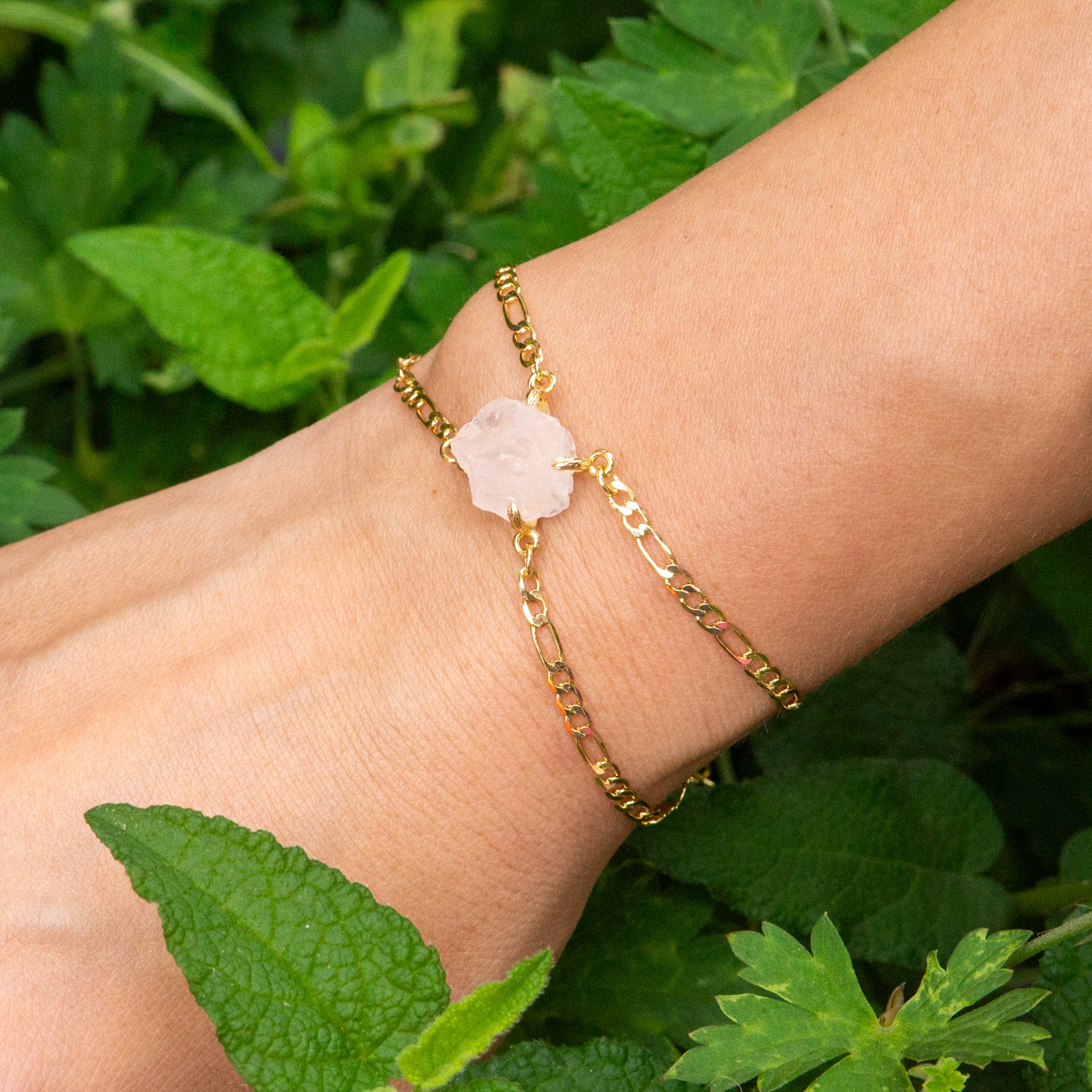 A delicate gold chain bracelet with a light pink rose-shaped stone charm, worn on a person's wrist against a background of green leaves. Elemental Rose Crystal Adjustable Bracelet  by wjy studio