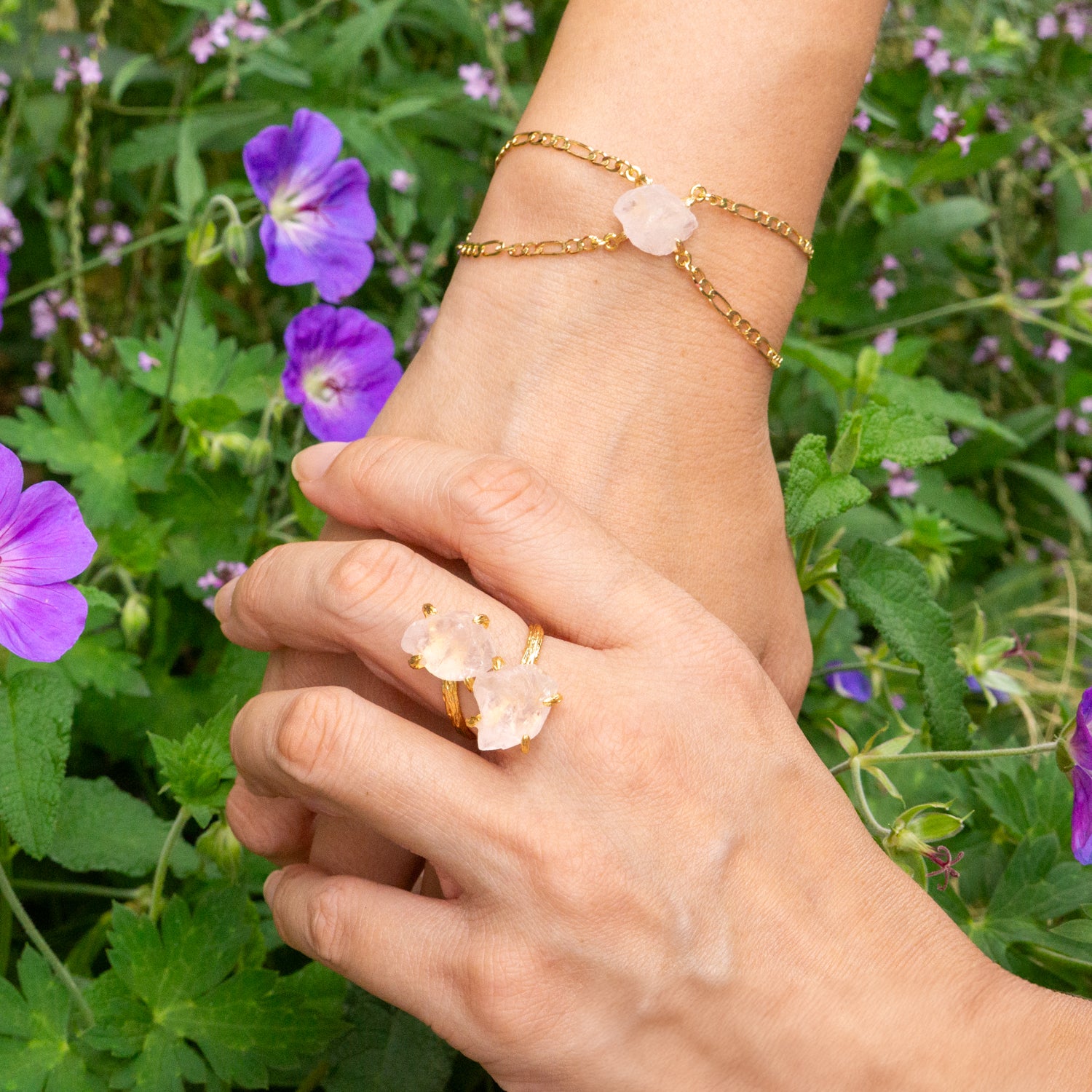 A close-up of two hands and a wrist wearing gold jewelry outdoors. The ring on the finger features a large pale pink rose quartz crystal, and the wrist has a delicate gold chain bracelet with a matching pink rose . quartz stone.Elemental Rose Crystal Adjustable Bracelet 
