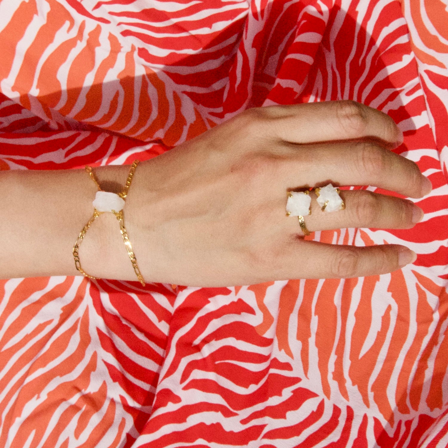 Elemental Rose Moonstone Bracelet by wjy studio featuring a central, rough-cut white crystal pendant  A close-up of a person's hand wearing two gold rings with white stone accents, resting on a bright coral and white zebra-print fabric background.
