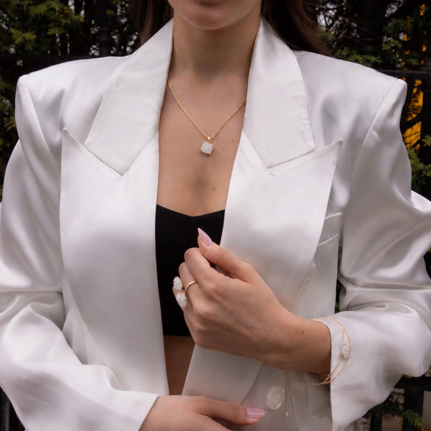 A close-up of a person wearing Elemental Rose Moonstone Bracelet by wjy studio featuring a central, rough-cut white crystal pendant, a white satin blazer over a black top, accessorized with a gold necklace, rings, and bracelets. The background is a blurred outdoor setting with green foliage.