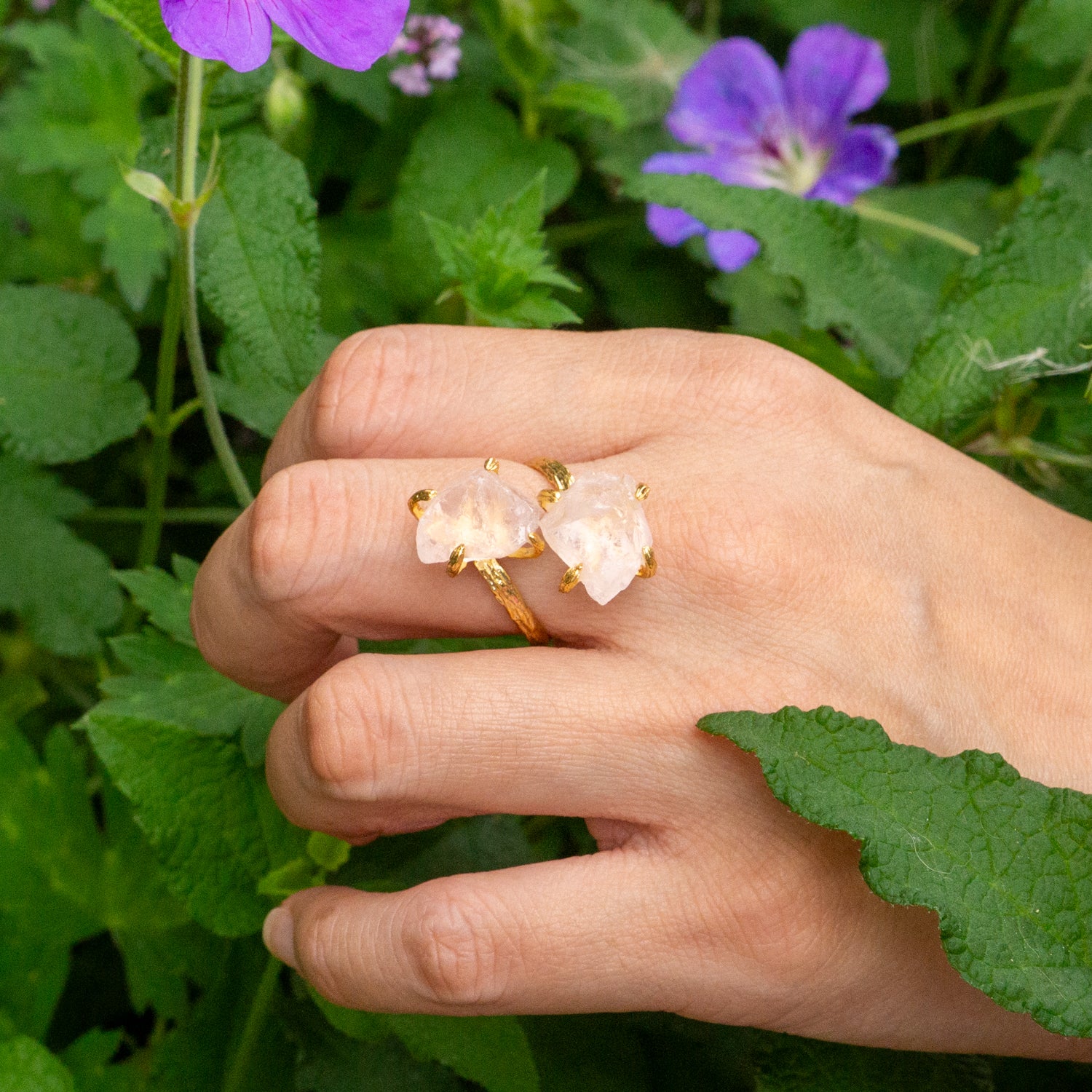 A close-up of a hand wearing A gold Elemental Rose Crystal Adjustable Ring by wjy studio  featuring a large pale pink rose quartz crystal, set against a background of green leaves and purple flowers.
