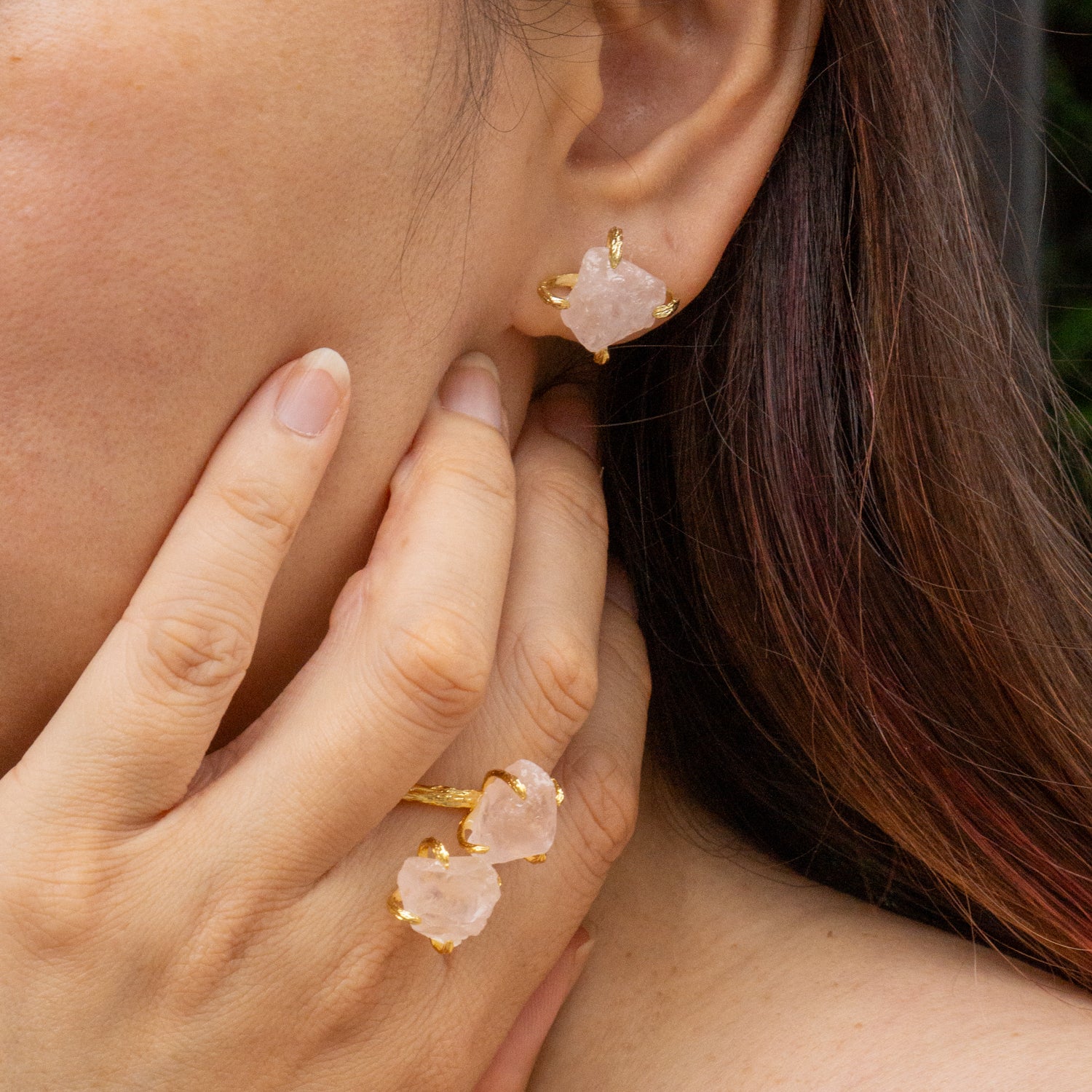 A close-up lifestyle shot of a person wearing gold-toned jewelry, featuring a gold Elemental Rose Crystal Adjustable Ring by wjy studio with two raw pink stone clusters and matching gold stud earrings.