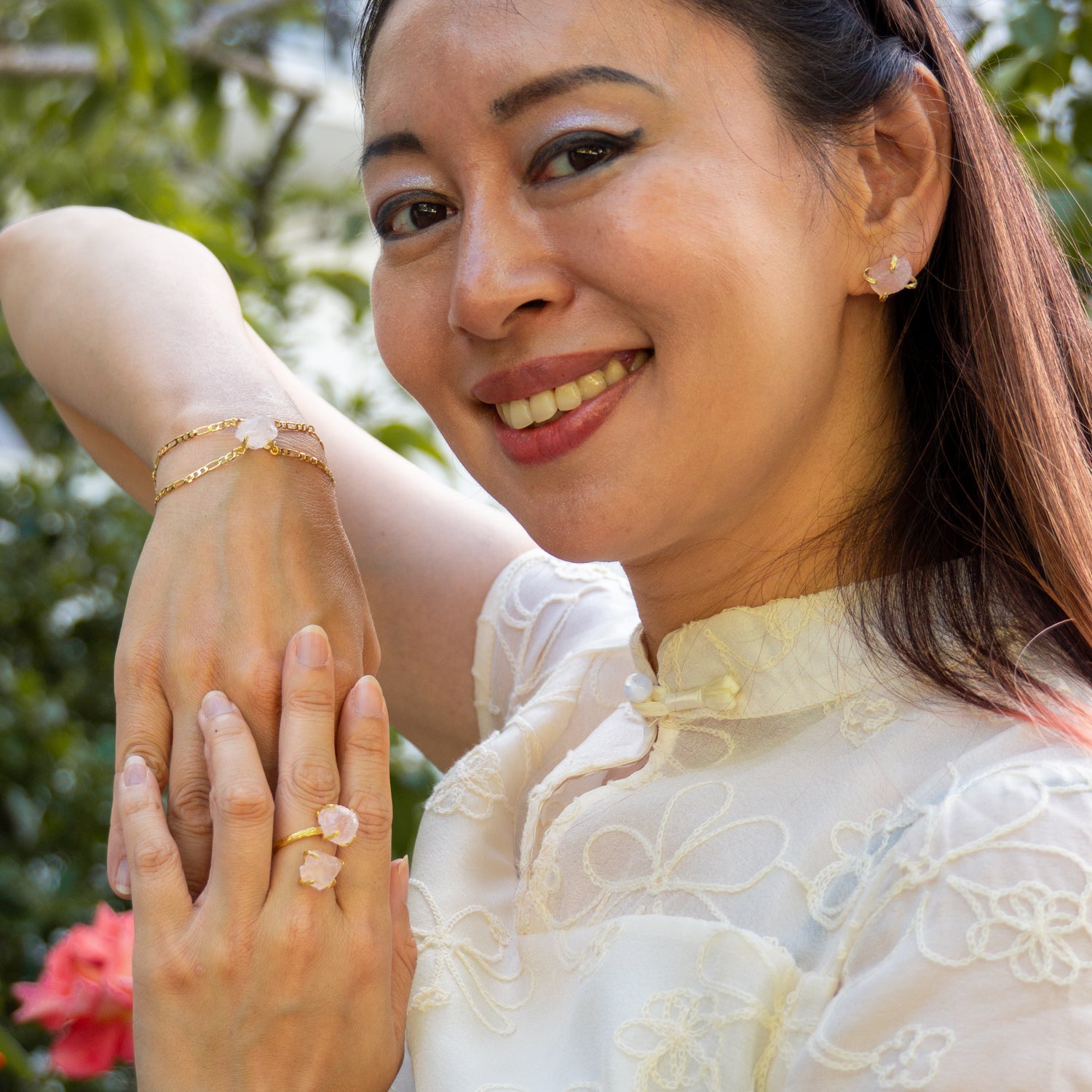 A close-up lifestyle shot of a woman wearing a delicate gold chain bracelet and a matching gold Elemental Rose Crystal Adjustable Ring by wjy studio with a pale pink stone, set against a soft-focus outdoor background of green foliage.