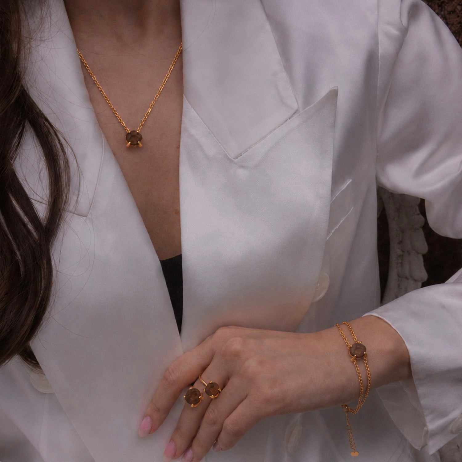 A close-up lifestyle shot of a model wearing a gold necklace with a dark brown geometric pendant, a matching gold bracelet, and two matching rings with round brown stones, styled with a white satin blazer.  Elemental Rose Tiger’s Eye Bracelet by wjy studio