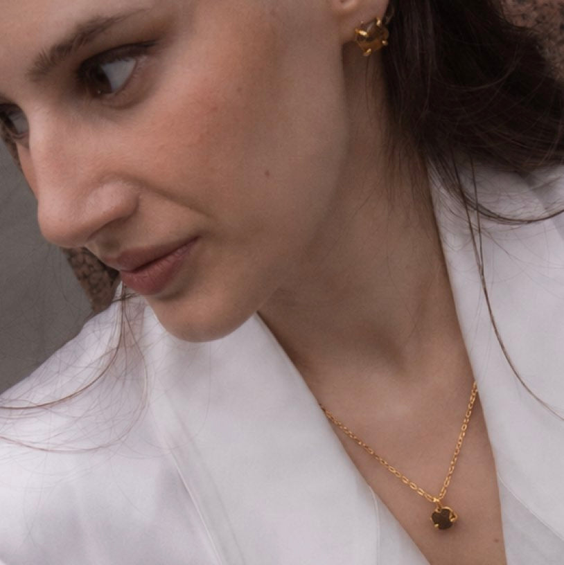 A close-up lifestyle shot of a woman wearing a delicate gold chain necklace with a small dark clover pendant, paired with a white blouse. from Elemental Birth Natural Tiger’s Eyes Stud Earrings by WJY Studio 