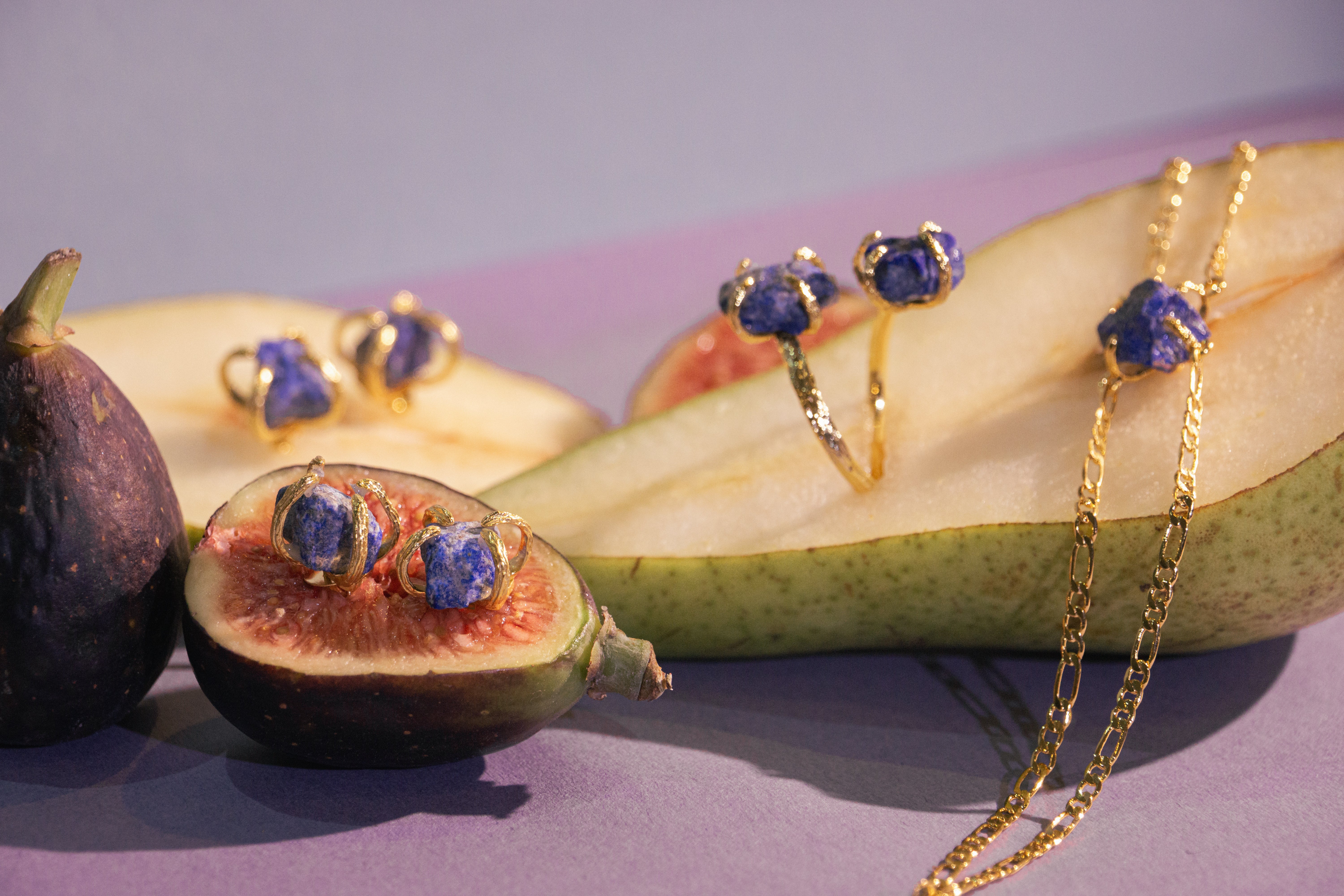 WJY Studio sapphire birthstone jewellery collection Gold jewelry pieces including rings, earrings, and a necklace with blue stone accents, styled on sliced pears and figs against a purple background. 